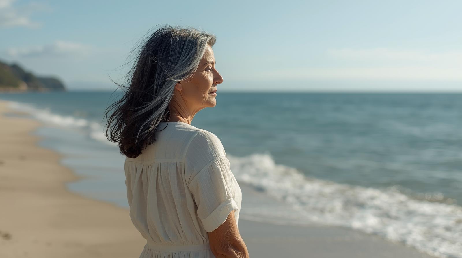 穏やかな海辺で、一人で風に吹かれながら遠くを見つめる、凛とした50代女性。